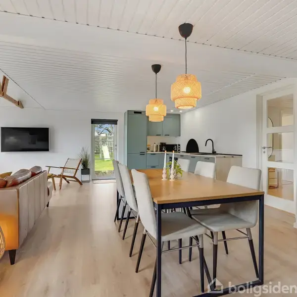 Spisestuebord med stole er centralt placeret; lamper hænger over bordet. I baggrunden ses en stue med sofa, brændeovn og vægmonteret tv. Glædende lys fra store vinduer.