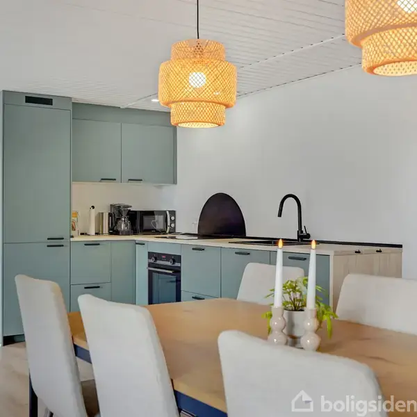 Køkken med lyseblå skabe og sorte greb ved siden af spisestue med lang træbord og hvide stole. To flettede lamper hænger over bordet. Store vinduer giver udsigt til have.
