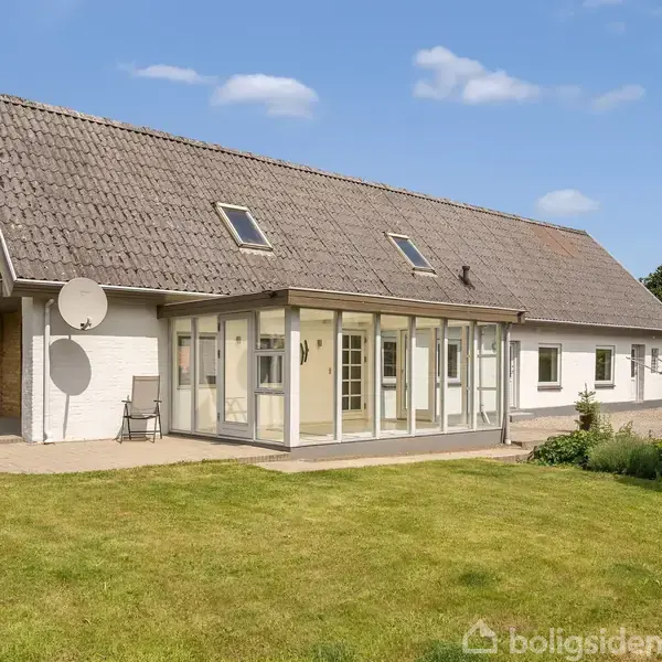 Et moderne hus med ovenlys og en stor glaseret veranda står i en grøn have. Huset har en grå tagkonstruktion, hvid facade, og der er en satellitantenne monteret på siden.
