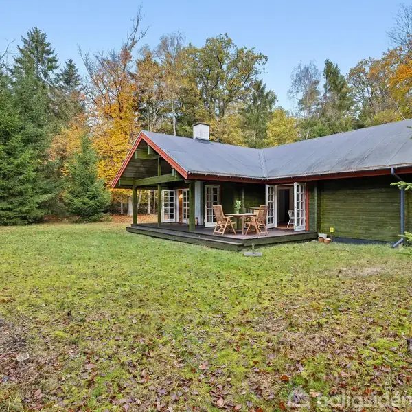 Et træhus står stille på en græsplæne i en skovomgivelse med tætte træer, omgivet af efterårsfarver. En terrasse med havemøbler ses foran huset.