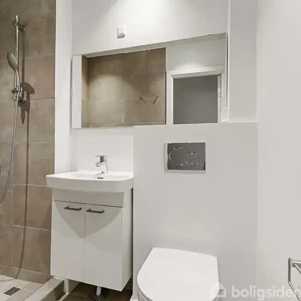 Brusekabine med håndbruser i moderne badeværelse ved siden af vaskeskab med spejl og hvidt toilet. Væggene er flisebelagte i neutrale farver.