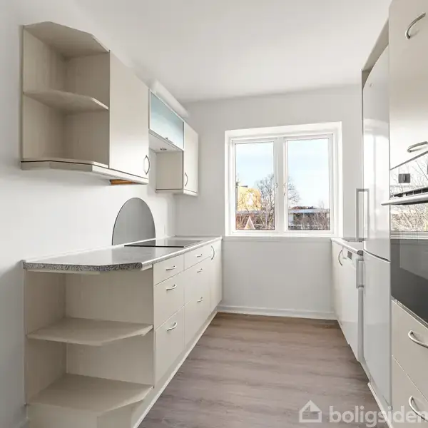 Et lyst køkken med hvide skabe og moderne apparater. Bordpladerne er grå, og der er et stort vindue, der giver naturligt lys. Trægulvet skaber en varm atmosfære.