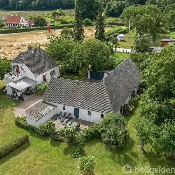 Hvidt hus og længe med grå tage ligger stille i en grøn have. Omkring dem ses træer, buske og marker. I baggrunden står et rødt hus ved en landevej.