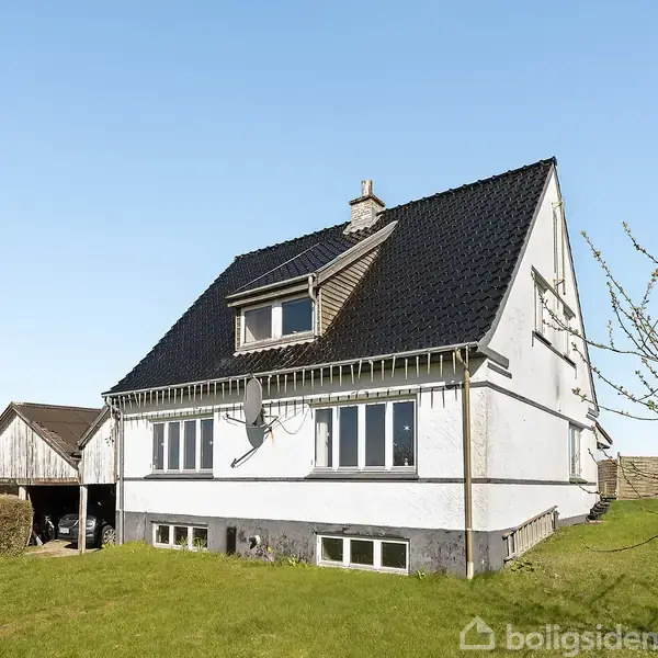Hus med sort tag står på en solrig græsplæne. Det har en kvist på taget og er omgivet af en hæk. Klar blå himmel i baggrunden.
