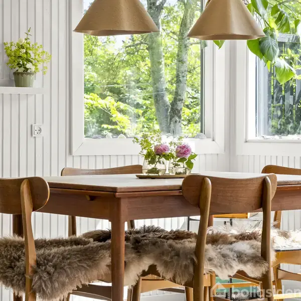 Et træbord med fire stole dækket af fåreskind er placeret i en lys stue. Bordet er dekoreret med blomster, og bagved er store vinduer med udsigt til grønne træer.