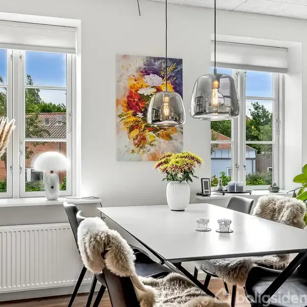 Spisebord med stole dækket af fåreskind står i et lyst spisestue med hængende lamper over. Dekorative planter og kunst pryder rummet, omgivet af store vinduer med udsigt til huse.