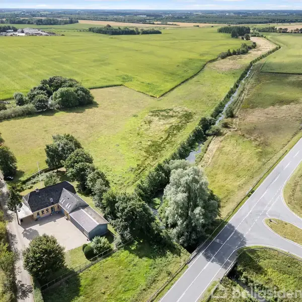 Et hus ligger ved en landevej omgivet af grønne marker og træer. En smal kanal løber langs vejens kant, der strækker sig gennem landskabet.
