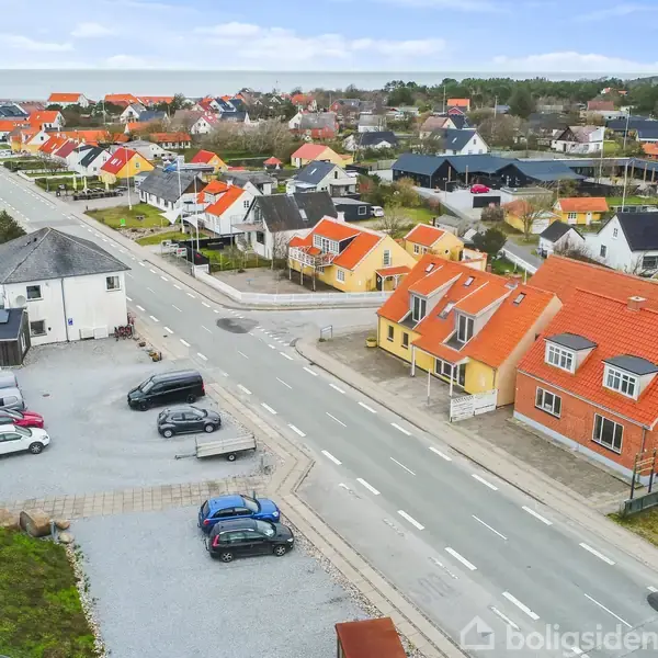 Biler holder parkeret i en grusparkeringsplads nær en vej med farverige huse. Huse med røde tage strækker sig langs vejen. Kysten ses i baggrunden.
