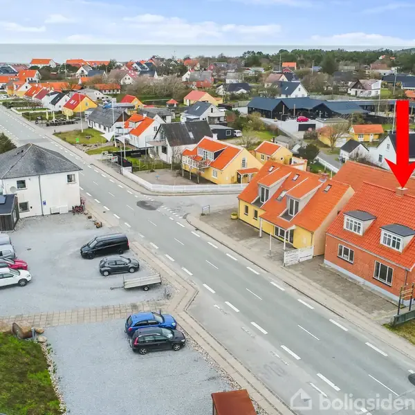 Rødt murstenshus peget ud med pil, stillet parallelt med vejen. Omgivet af andre huse med farverige tage og en parkeringsplads med flere biler. Kysten anes i baggrunden.