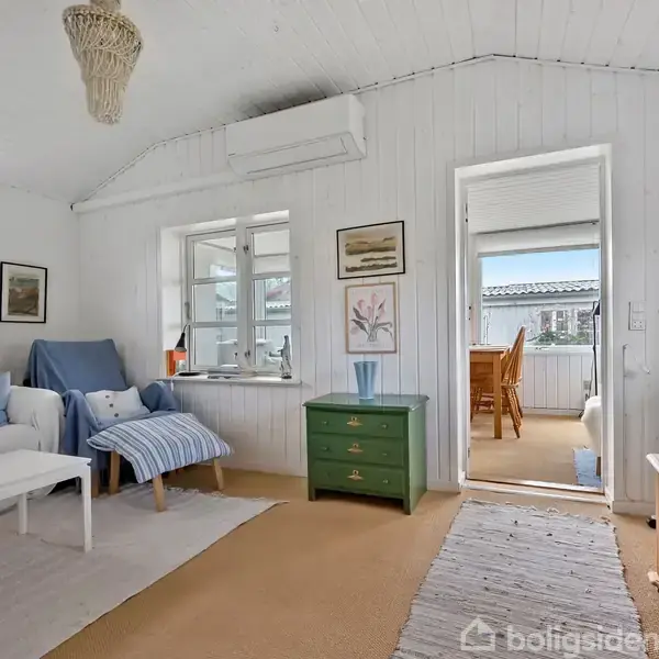 Stue med hvidt træloft og vægge. Sofa med blå betræk ved siden af hvidt bord. Grøn kommode med vase. Vinduer giver udsigt til solrig veranda med stole. Rug på gulvet.