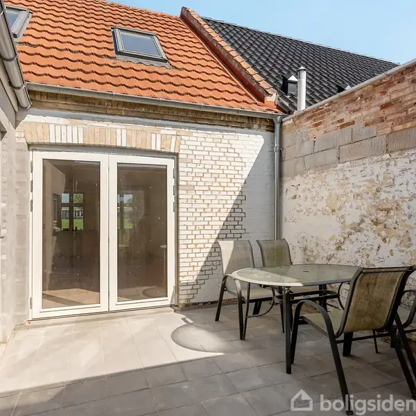 Terrassemøbler står på en flisebelagt gårdhave med tre lænestole omkring et rundt bord. Døre med glasruder fører ind i huset, omgivet af hvidmalede og rå murstensvægge.