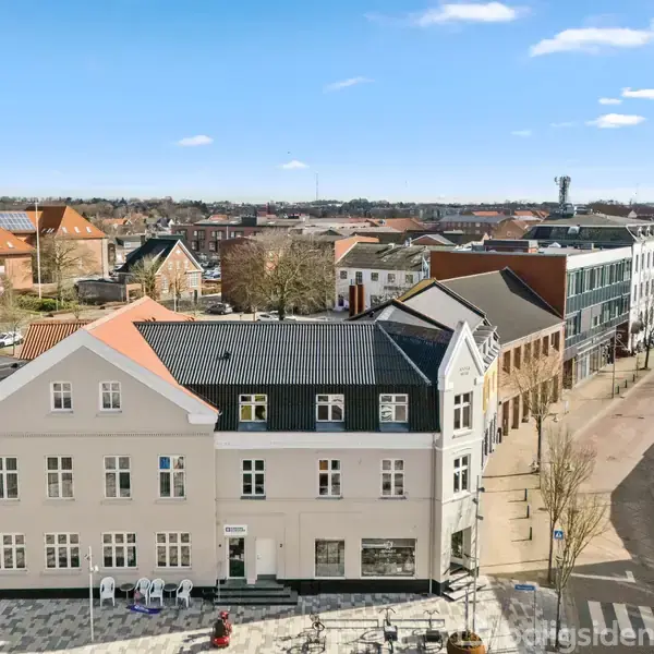 Et hvidt, tre-etagers hus med skråt tag står ved en byvej. Solen skinner, og flere huse strækker sig langs vejen i baggrunden.