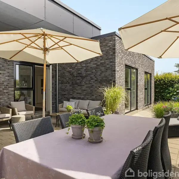 Parasol dækker bord med stole på terrasse, omgivet af moderne murstensbygning og grønne planter. Kurvemøbler og potteplanter skaber en indbydende udendørs opholdszone.