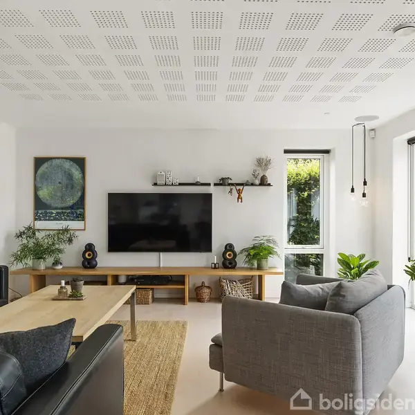 Stue med sort lædersofa og grå lænestol, centreret omkring et sofabord og en stor fladskærms-TV. Store vinduer giver udsigt til en grøn have.
