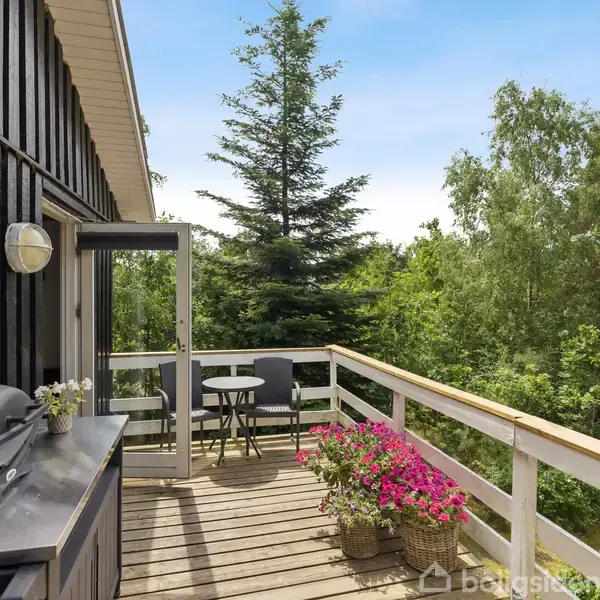 Terrasse med grill og blomster. En sortmalet trævæg til venstre har en lampe. Et lille bord og to stole er placeret ved rækværket, omgivet af grønt løv og høje træer.