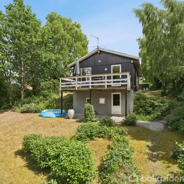 Et sorte træhus står midt i en frodig have med høje træer. Huset har en altan, og der er en lille pool i haven.