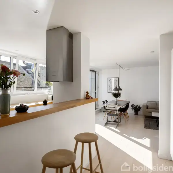 Et moderne køkken åbner op til en lys stue. To barstole står ved en træbar. En vase med blomster står på bordet. Store vinduer giver meget lys.