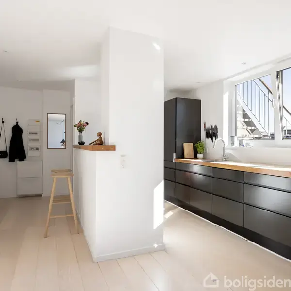 Et moderne køkken med sorte skabe strækker sig langs en væg, der har et stort vindue. Til højre er en hvid reol og en knagerække i en lys gang.
