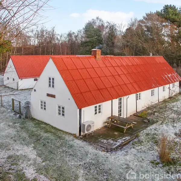 Hvid bygning med rødt tag, står stille omgivet af let snedækket græs og træer i baggrunden. Bænkesæt foran huset, der er et typisk landhus i naturligt skovområde.