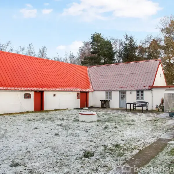 Huset har hvid facade og rødt tag, står i en have dækket af let sne. En rund brønd står i forgrunden, mens træer omkranser ejendommen i baggrunden.