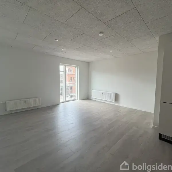 Et stort, tomt værelse med lyse trægulve og hvide vægge. Et vindue til venstre giver udsigt til en bygning udenfor. To radiatorer er monteret ved væggene.