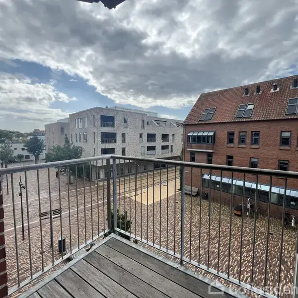 Balkon med metalgelænder strækker sig ud over en brolagt plads. Omgivet af moderne og traditionelle bygninger under en skyet himmel.