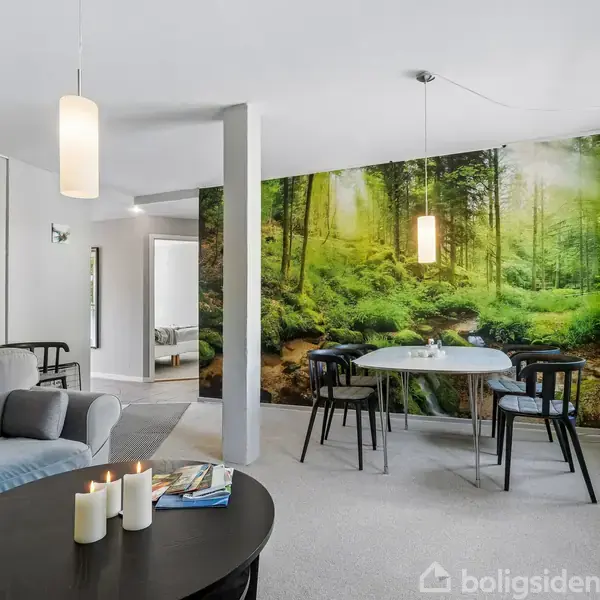 Stue med grå sofa og rundt sofabord med stearinlys. Baggrunden er et stort fototapet af en frodig skov. Rundt spisebord med stole står ved siden af et vindue med grønne gardiner. Tre små billeder hænger på væggen.