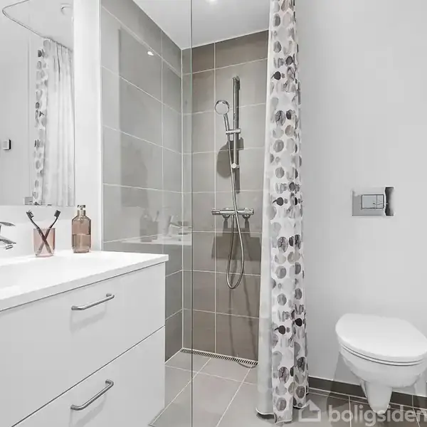 Badeværelse med hvid vask og skab, bruser i glasafskærmning med mønstret forhæng, og væghængt toilet. Miljøet er lyst og minimalistisk.