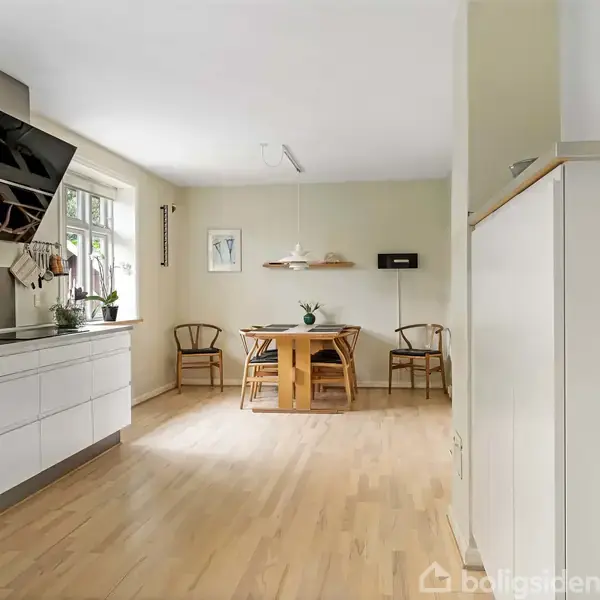 Køkken-alrum med hvidt køkkenbord langs venstre væg. Træspisebord med tre stole står ved siden af et vindue og vægdekorationer. Lys trægulv og beige vægge.