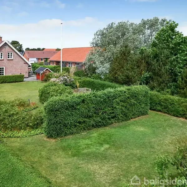 Et rødt murstenshus med hvidt tag står i en velplejet have omkranset af hække. Græsplænen er stor, og der er flere træer og buske i landskabet.
