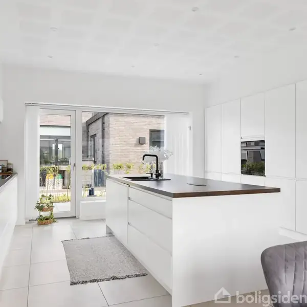 Et moderne køkken med hvidt interiør, øen har sort vandhane, der leder til glasdøre med udsigt til en have. Gråt gulvtæppe og stol til højre.