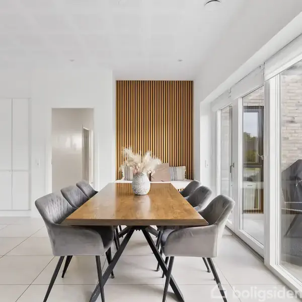 Et langt spisebord er omgivet af seks grå stole i en moderne spisestue. Store vinduer på højre side giver udsigt til en terrasse. En hvid vase står på bordet.