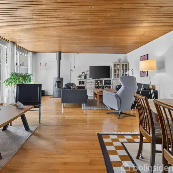 Stue med træloft, sofa og lænestole omkring et sofabord, brændeovn i baggrunden, og spisebord i forgrunden. Store vinduer med udsigt til grønne planter udenfor.