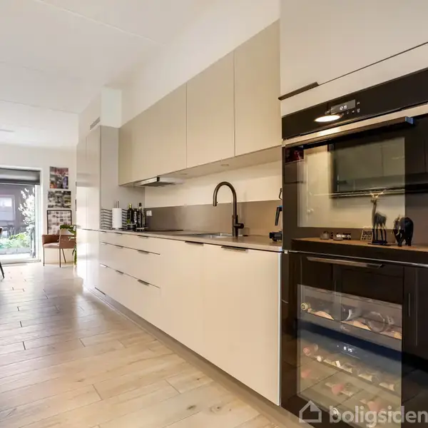 Et moderne køkken med sorte og lysegrå skabe indeholder en sort ovn og indbygget mikrobølgeovn. Køkkenet fører til en spisestue med træstole, et bord og glasdør mod haven.