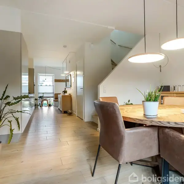 Rundt spisebord med brune stole står i stue med lyse trægulve. På bordet er planter og lysestager. Væggene har billeder og planter. I baggrunden ses køkken og trapper.