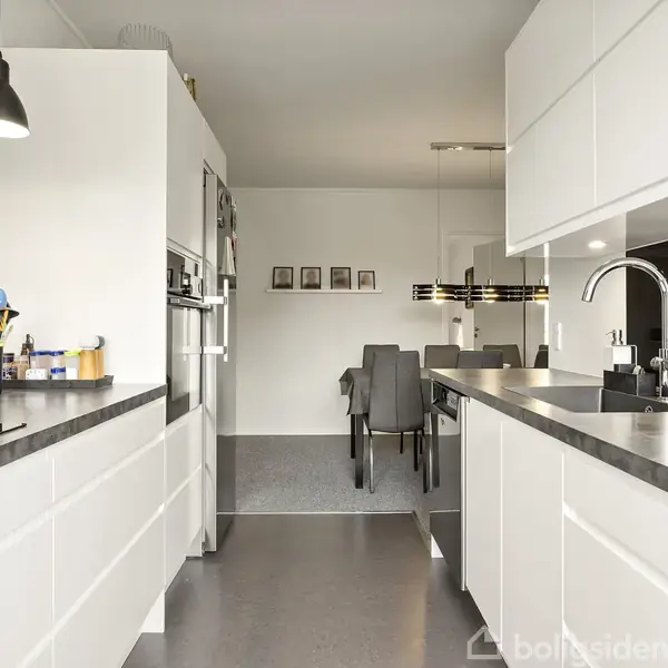 Køkken med hvide skabe, sort bordplade og dobbelte vaske; står ved siden af spiseplads med grå stole og moderne lysarmatur. Fire billeder hænger på væggen i baggrunden.