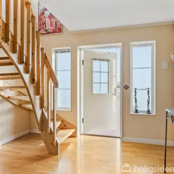 Trappe af træ fører opad i en lys entre med lyse vægge. Til højre står et konsolbord, mens dør og vinduer ses i baggrunden.