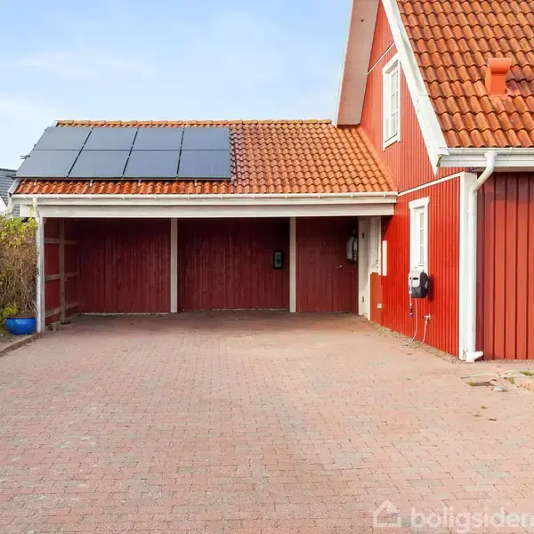 Rødt hus med solpaneler på taget. Carporten er åben og flankeret af en murstensbelagt indkørsel. Huset har hvide vinduesrammer og ligger i et forstadsmiljø omgivet af buske.