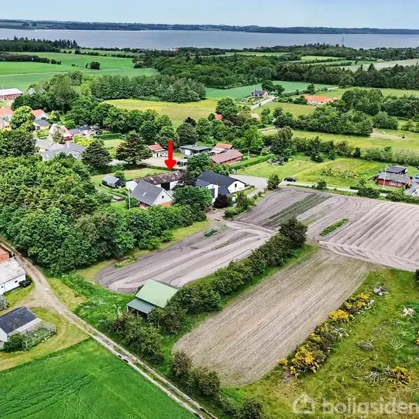 Røde tage ligger blandt grønne marker og bygninger, en rød pil peger mod et hus. Landskabet omfatter skove, marker og en fjord i baggrunden.