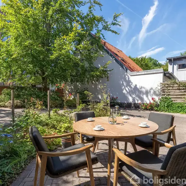 Et rundt træbord står omgivet af stole i en hyggelig gårdhave. Tallerkener og en karaffel er på bordet. Omkring er der blomsterbede, træer og en hvid mur.
