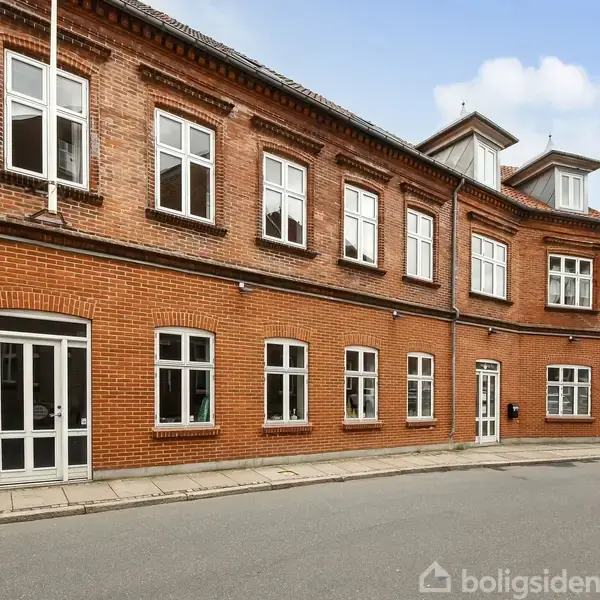 En to-etagers murstensbygning med hvide vinduer står langs en stille gade. Bygningen har flere vinduer og en hoveddør, omgivet af blå himmel med få skyer.