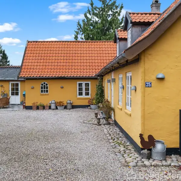 Gul bygning med rødt tegltag står stille på en gårdsplads med grus og planter. Blå husnummerplade viser "25", omgivet af grønne træer og blå himmel med skyer.