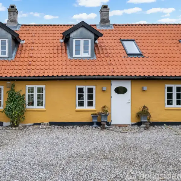 Gult hus med rødt tegltag står stille på grusplads, omgivet af planter i potter og en blå himmel med få skyer. To loftsvinduer og tre skorstene ses på taget.