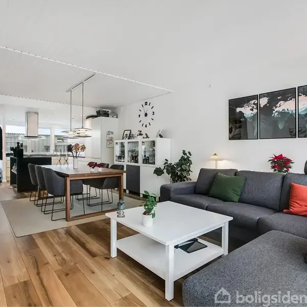 En stue med en grå hjørnesofa og sofabord. Spisebord med stole i baggrunden. Vægdekoration og planter skaber et moderne hjemmemiljø. Kækken kan ses i det fjerne.