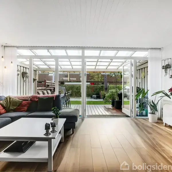 Sofa står i moderne stue, dekoreret med puder, ved siden af hvidt sofabord. Stuen åbner ud mod en terrasse med glasloft, omgivet af planter og græsplæne, med TV på væggen.