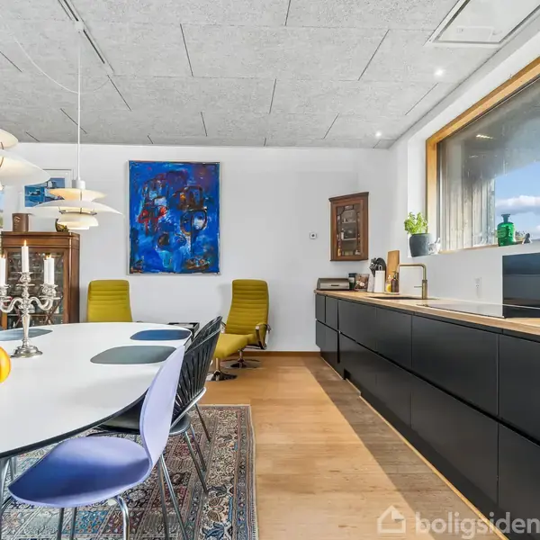 Et moderne køkkenalrum indeholder et langt, sort køkkenelement med store vinduer. Et ovalt spisebord med blå stole og en gul kande står i fokus. Kunst pryder væggene.