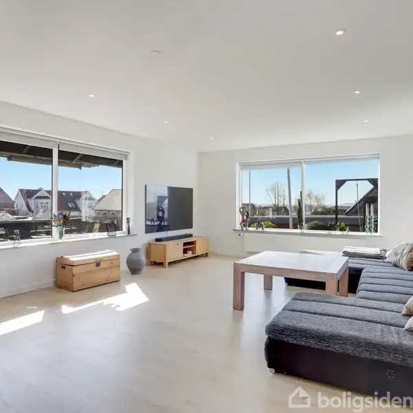 Stue med stor grå sofa, placeret ved et stort bord, i et lyst rum med trægulv og store vinduer, der giver udsigt til et boligområde.