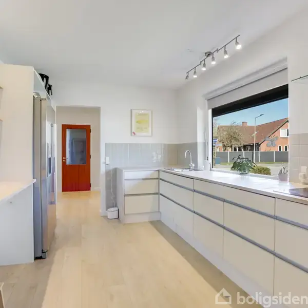 Køkken med hvide skabe langs væggen, overskabe og indbyggede apparater. Der er et stort vindue med udsigt til en forstadsvej. Lys træfarvet gulv og hvidmalede vægge.
