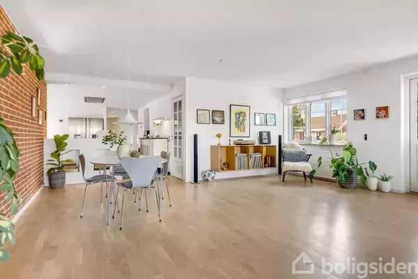 Et lyst rum med trægulv, spisebord og stole. Grønne planter står ved en murstensvæg. Et hjørne har reol, lænestol og billeder på væggen. Vinduer slipper lys ind.