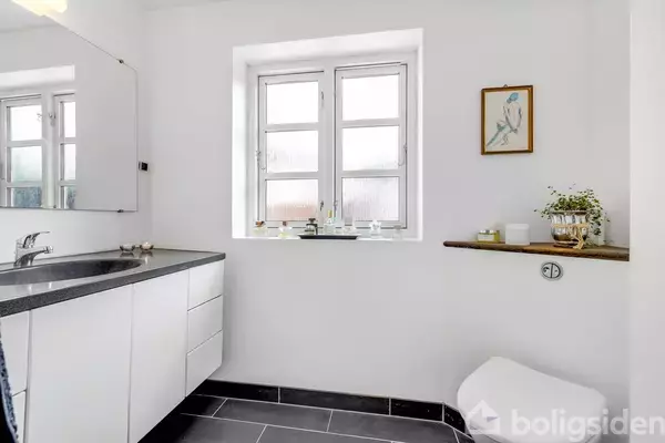 Badeværelse med hvidt skab og sort bordplade, vask, og spejl til venstre. Toalett til højre nær et vindue med planter, på en hylde hænger et billede.